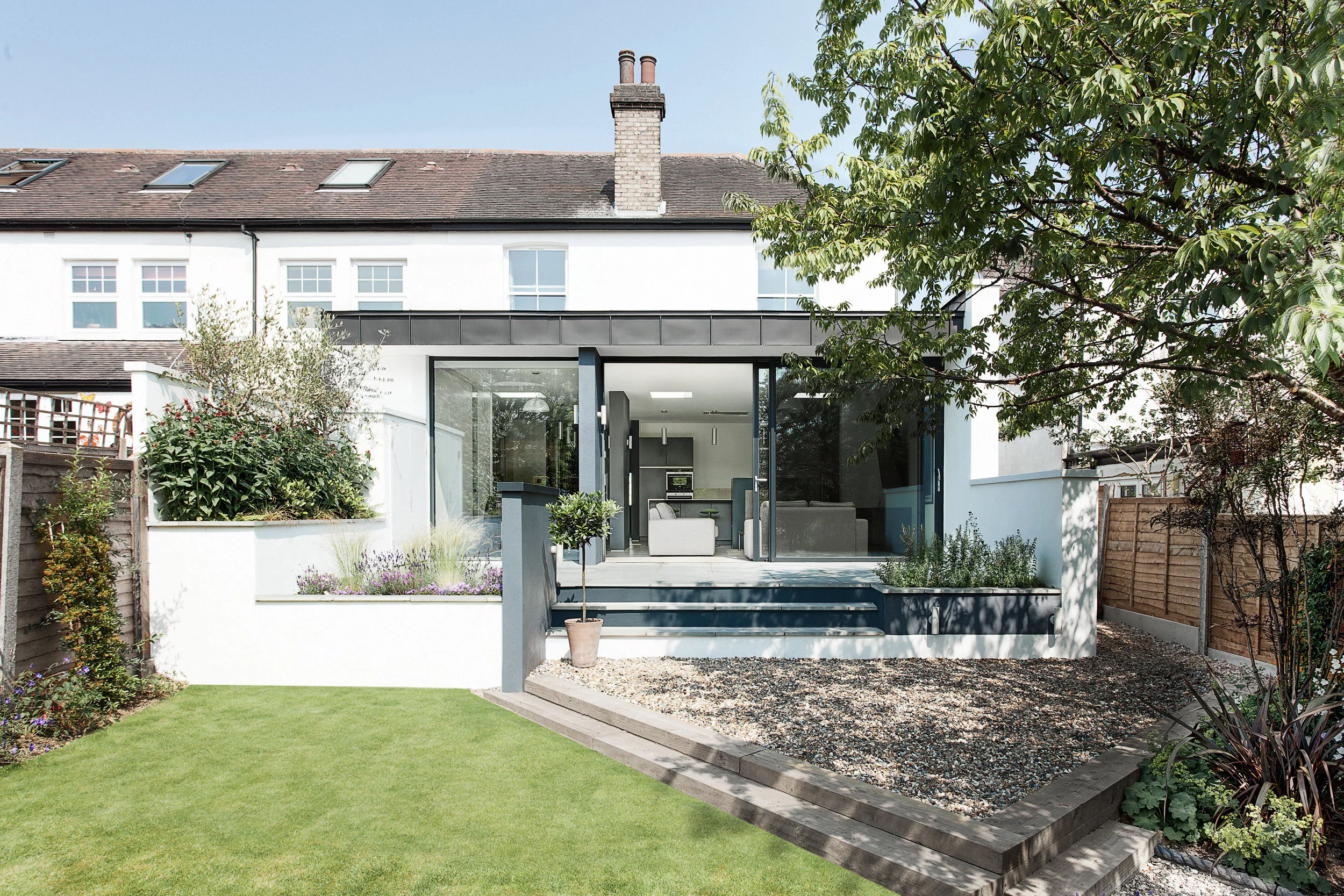 Elm Court is a single-storey modern extension to a three bedroom house in North London. The finished property retains its humble street appearance, yet is completely transformed from its previous gloomy atmosphere, with the new modern extension creating a light, airy and open living environment. The recently retired owners sought to adapt the existing small and cramped property into their perfect home. The brief was to design a single storey contemporary extension at the rear of the existing property, linking in with the original, whilst making minimal physical changes. The open-plan extension was to comprise of a new kitchen, dining/living space, utility room, cinema room, WC and office area; with the aim to bring views of the garden and more light into the property. “At AR Design Studio we are particularly interested in the relationships between inside and outside spaces, between the man-made and nature,” states lead architect Mike Ford. “The owners could see the potential to completely revive the feel and functionality of this home using the tight site as the parameter for an interesting design approach. The result completely transforms the feel and atmosphere of the existing property.” The architectural concept is rooted in uniting the house with the garden and positively affecting the interior feel of the original property, whilst making minimal physical changes. The solution was to have a wall running through the entire home. This physical wall acts as an axis of movement, guiding you through the house, from old to new. It also draws the eye through the space and out to the garden providing a physical inside/outside link. The wall breaks down as it dissects the contemporary extension, defining smaller, more intimate spaces, whilst still retaining the feeling of togetherness evident in open-plan living. As the wall reaches into the garden it projects through the large glazed doors, which can be opened completely. These allow sunlight to flood through the open-plan space and filter down gradually into the original property, creating beautiful shards of light. A flush threshold allows the tiled floor finish of the interior to continue out onto the patio, creating a seamless link between inside and outside.