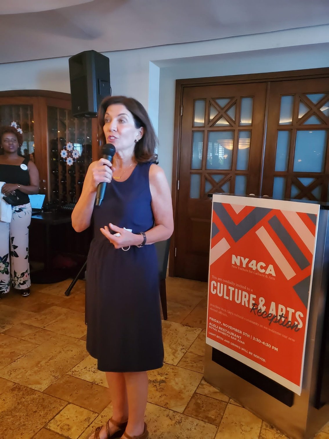  Kathy Hochul speaks into a microphone standing in front of a podium that features a “NY4CA Culture &amp; Arts reception” banner 