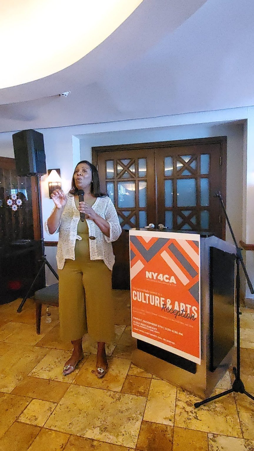  Tish James speaks into a microphone standing in front of a podium that features a banner that reads “NY4CA Culture &amp; Arts reception”  