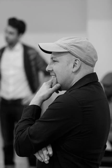 A man wearing a baseball cap and black shirt, smiling with his hand on his chin, in a room with another person in the background.