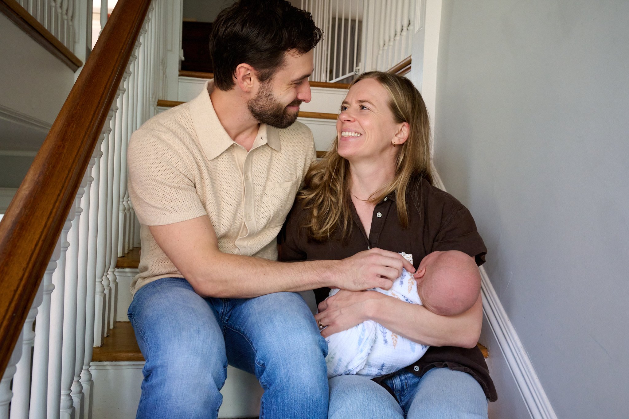 Mom and dad smile lovingly at each other with newborn baby on staircase of their West Hartford, CT home by family photographer Pamela Einarsen, 