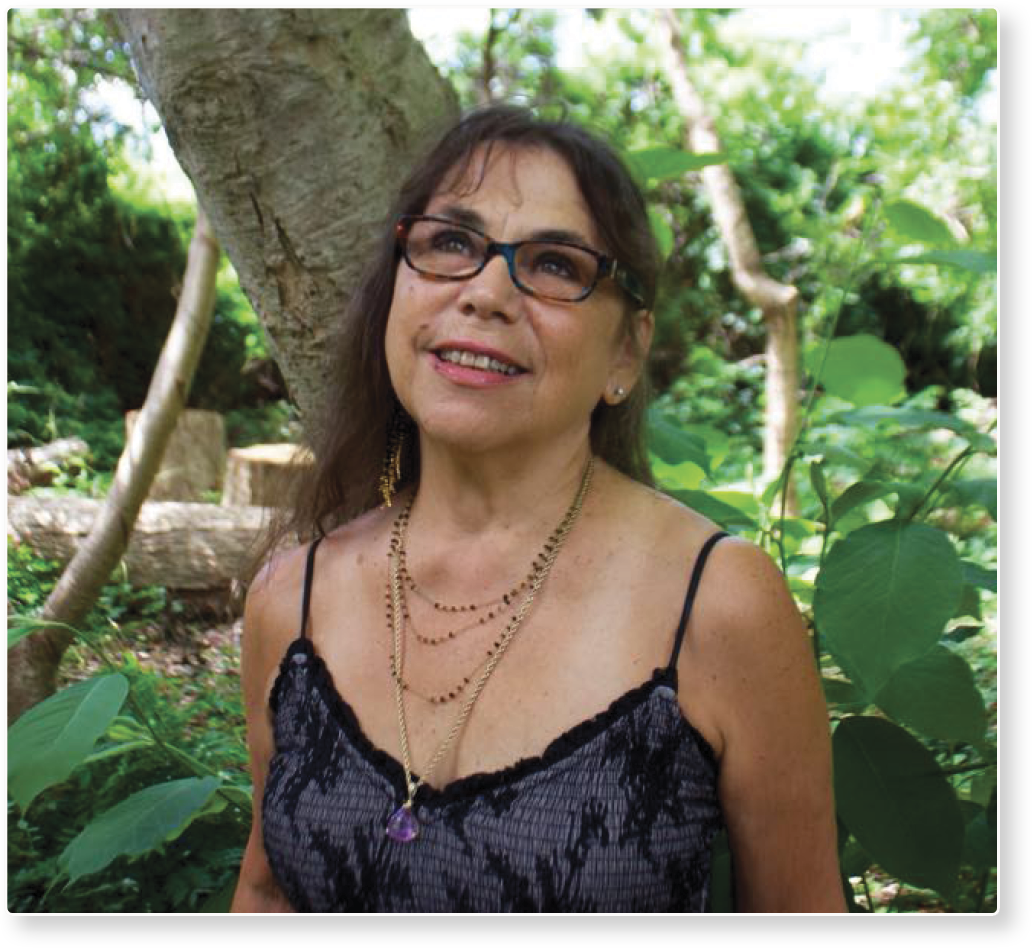 A woman with glasses and multiple necklaces standing outdoors in a lush, green forest, smiling and looking slightly upward.