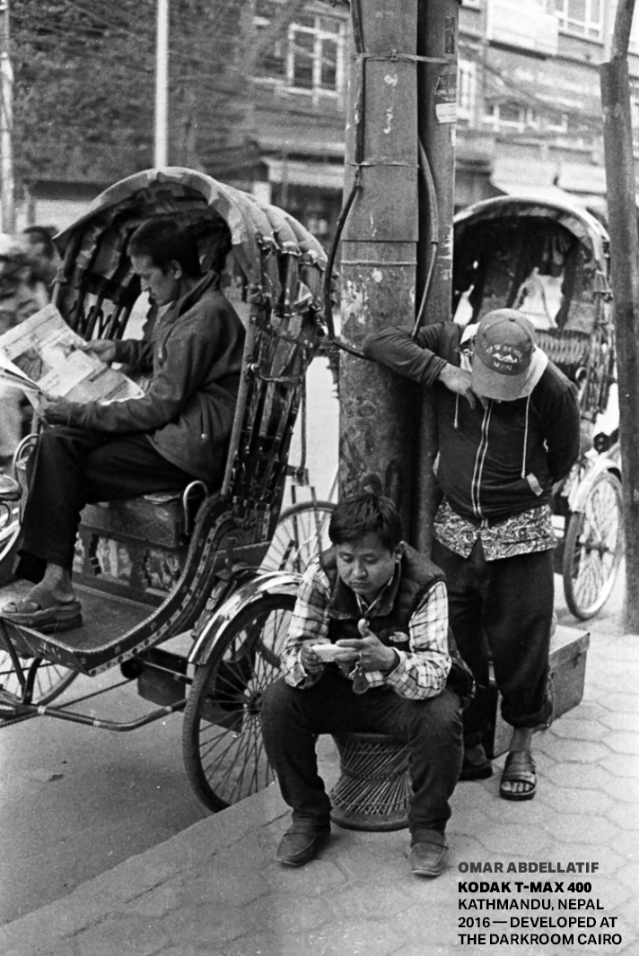 Three boys near a pole on a city street; one boy is sitting on a small stool looking at a mobile device, another boy leans against the pole looking down, and the third boy is seated on the curb reading a newspaper with a rickshaw behind them.