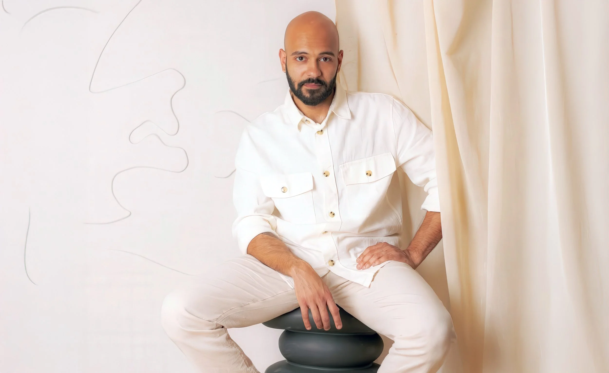A bald man with a beard sitting on a black stool, wearing an all-white outfit, in front of a plain light-colored background with minimal abstract line art.
