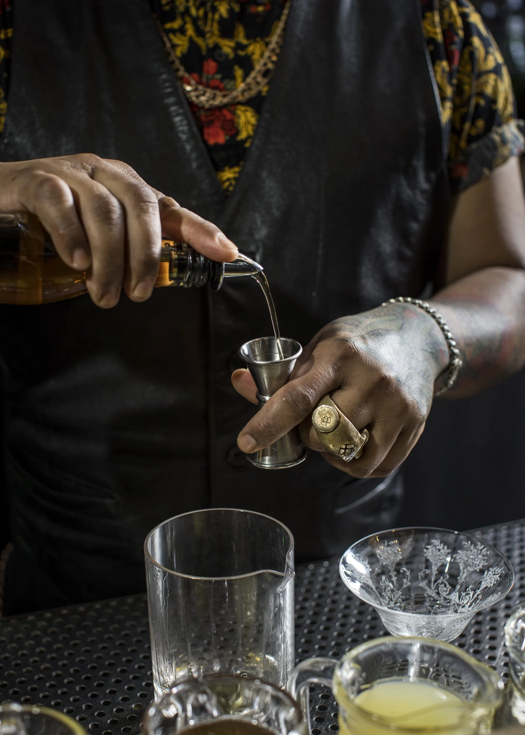 Person pouring a liquid from a bottle into a small metal jigger, with various glassware and ingredients on a bar counter.