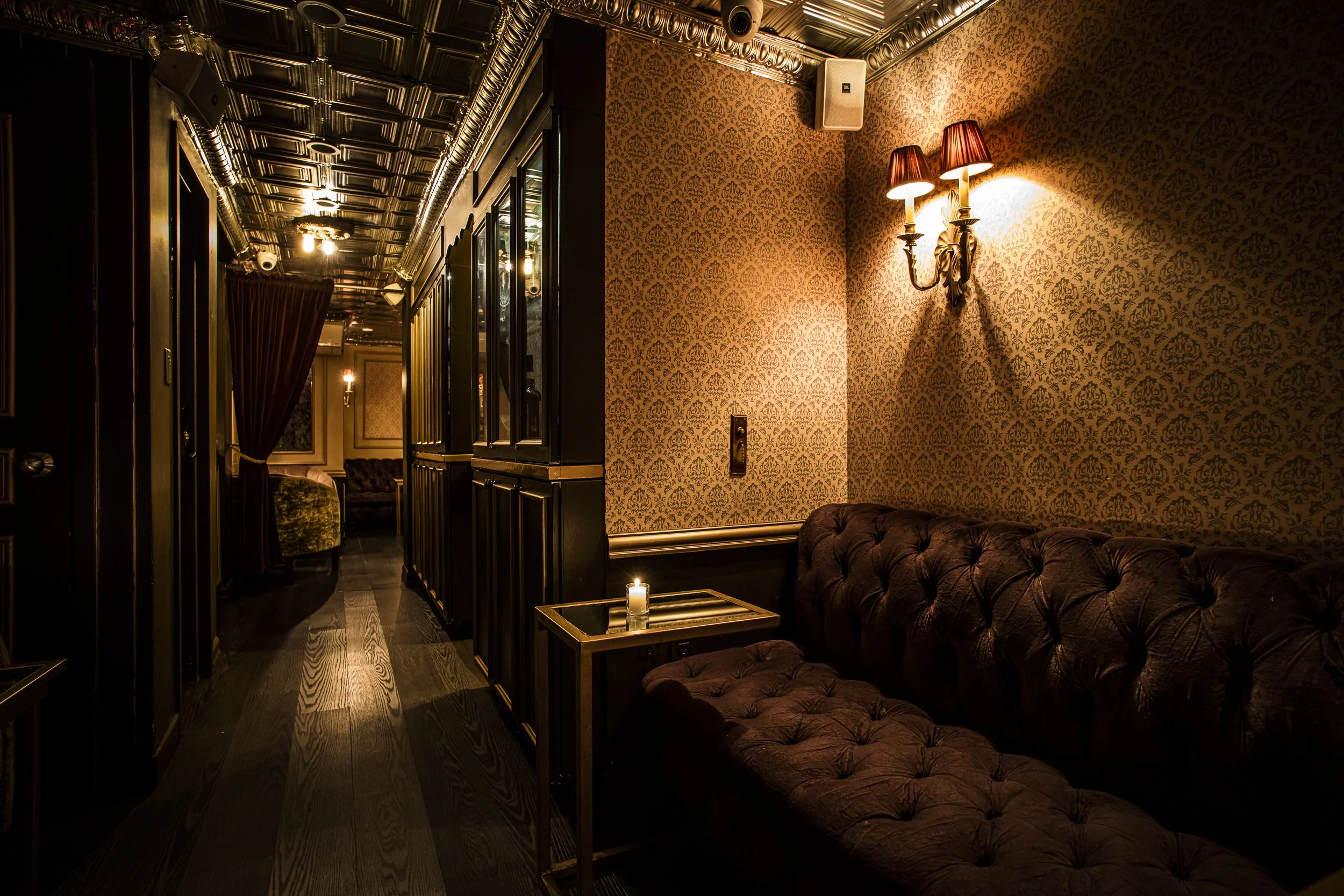 A dimly lit, elegant lounge with vintage wallpaper, a tufted leather sofa, a small glass-topped side table with a candle, ornate wall sconces, and a dark, polished wooden floor. The ceiling has decorative tin tiles, and there's a hallway with mirrors