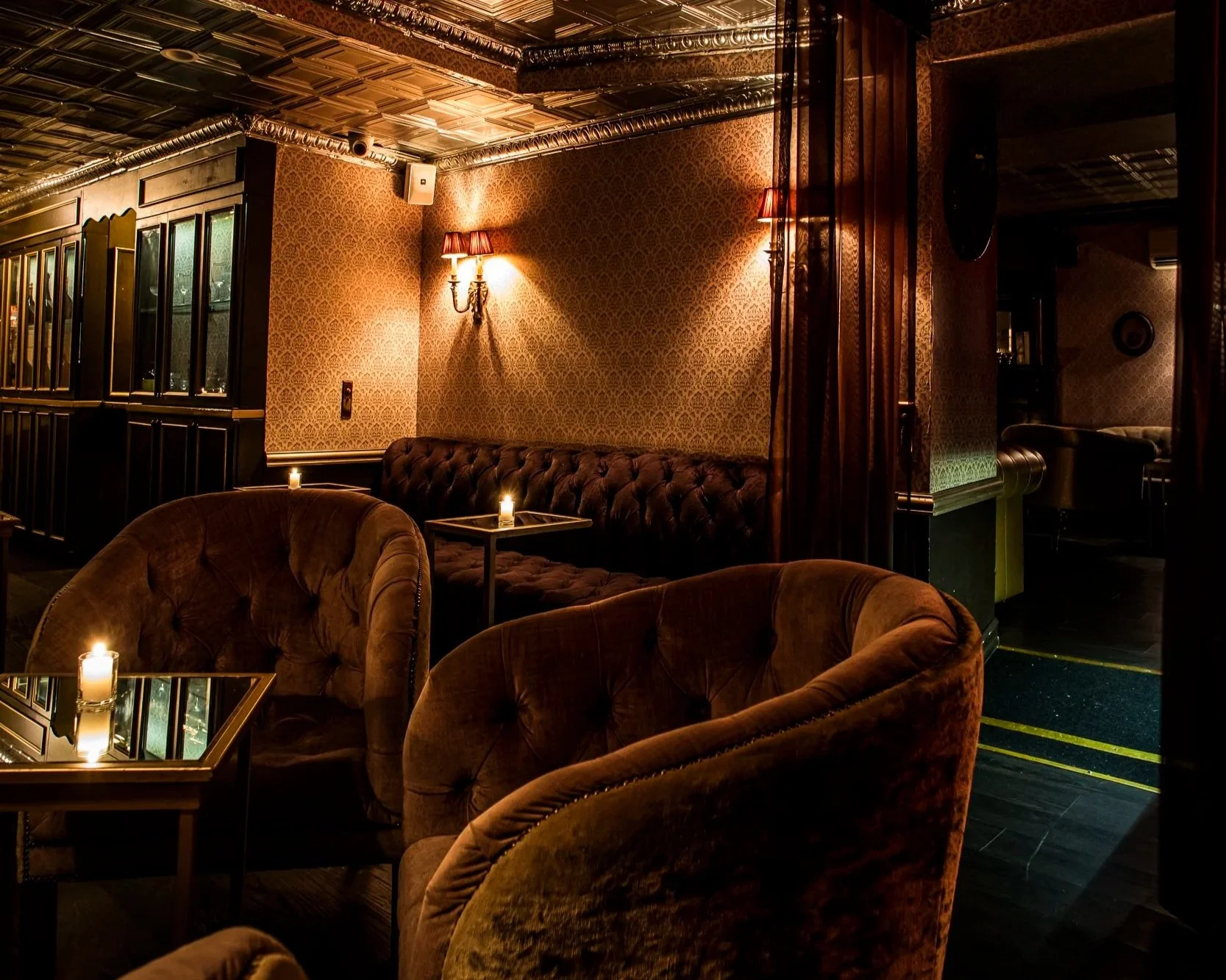 A dimly lit lounge area with plush velvet armchairs and a tufted sofa, candles on tables, ornate wallpaper, wall sconces with red lampshades, and reflections of other seating areas.