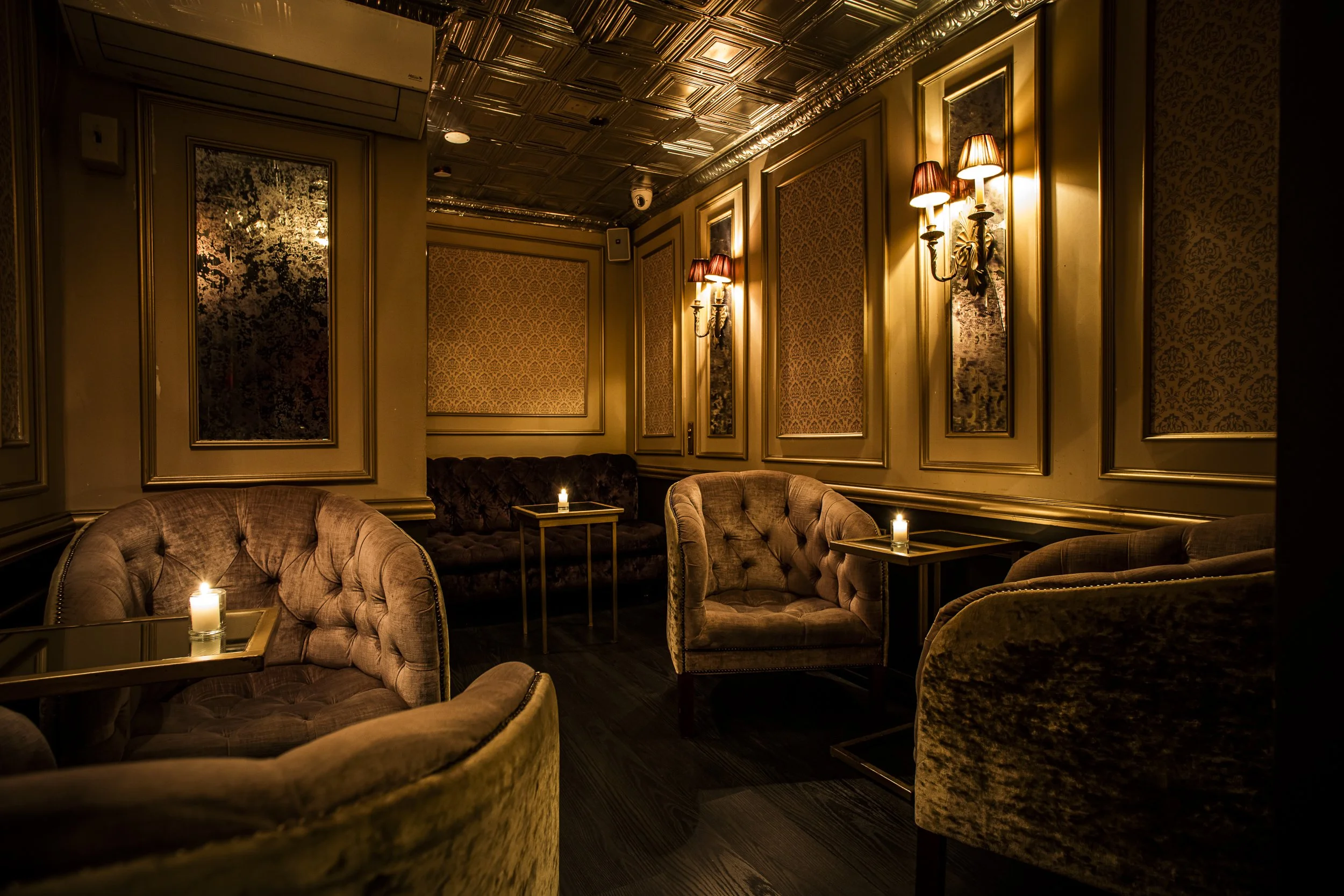 A luxurious, dimly lit lounge with plush velvet armchairs, small tables with lit candles, wall-mounted ornate lamps, framed decorative wallpaper, and a decorative tin ceiling.