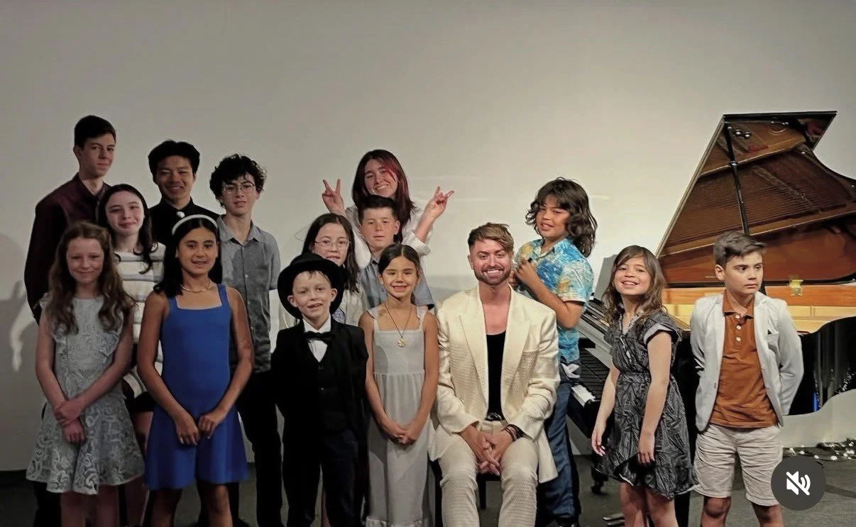 A group of children and two adults posing in front of a grand piano on a stage, some making peace signs and all smiling.