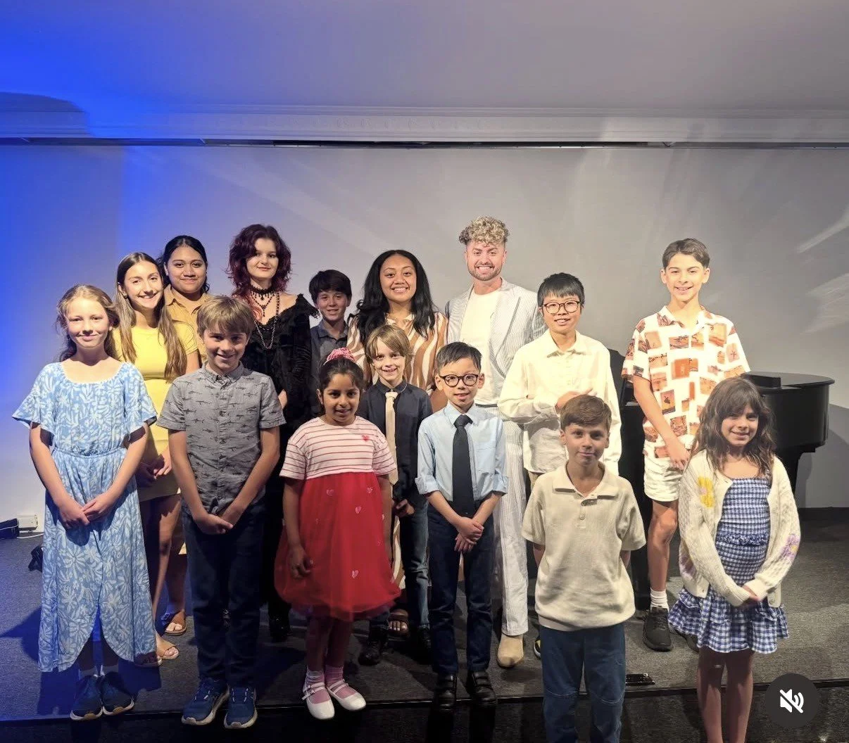 Group of children and adults posing for a photo indoors, some standing and some sitting, with a gray wall background and a black piano to the right.