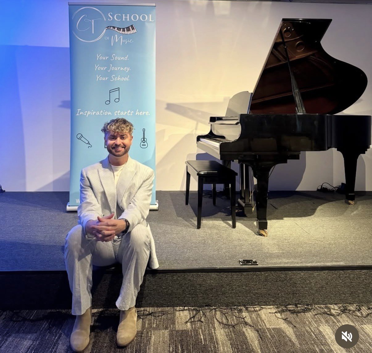 A man in a light-colored suit sitting on the edge of a small stage in front of a blue banner that reads 'School of Music' and shows musical notes, a microphone, and guitars. To his right, there is a black grand piano with an open lid and a matching bench.