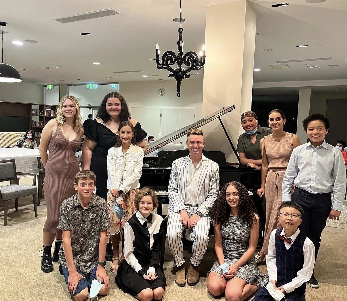 A group of people, including children and adults, gathered around a black grand piano in a well-lit room. Most are smiling, and some are wearing masks pulled down. There is a chandelier hanging from the ceiling and a bookshelf in the background.