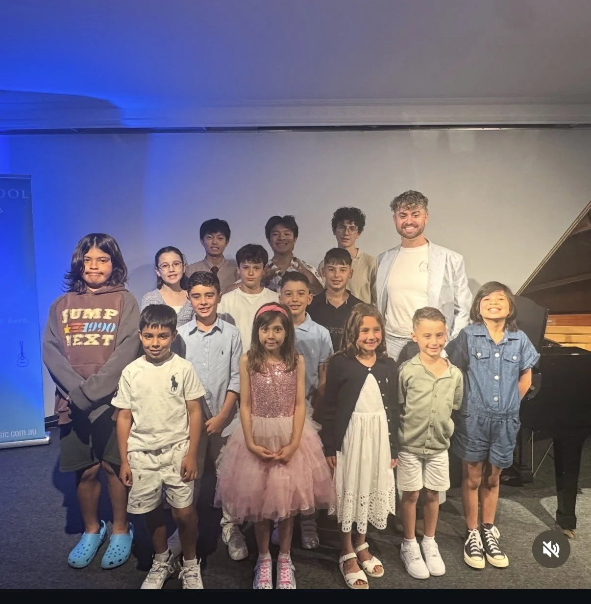 Group of children and an adult man posing together on stage with a piano in the background, some children are smiling and dressed in casual and semi-formal clothes.