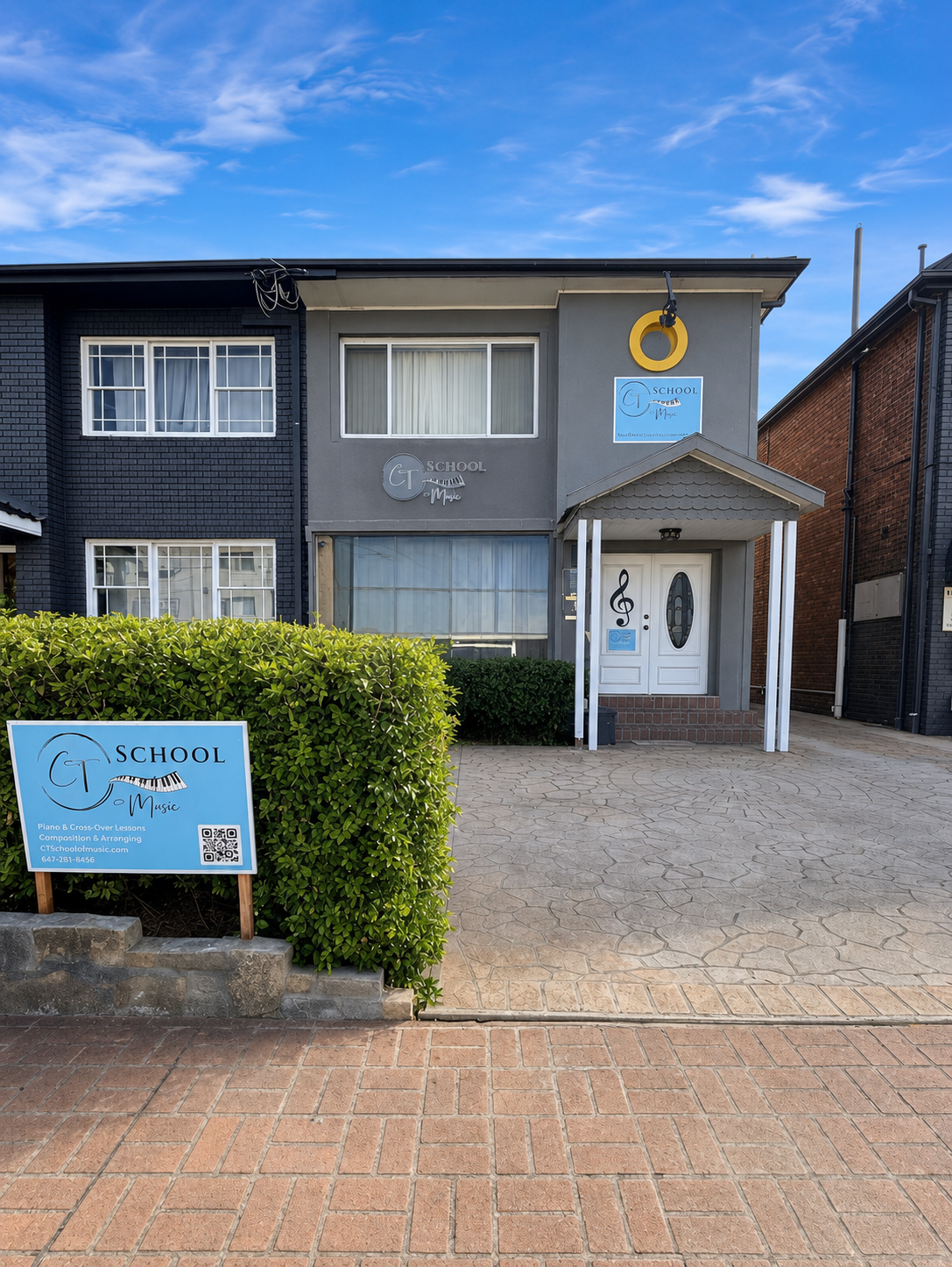 Exterior of a gray two-story music school building with piano and musical note signs, a blue and white school sign, and a large yellow ring sign above the entrance. A sidewalk and green hedge are in the foreground.