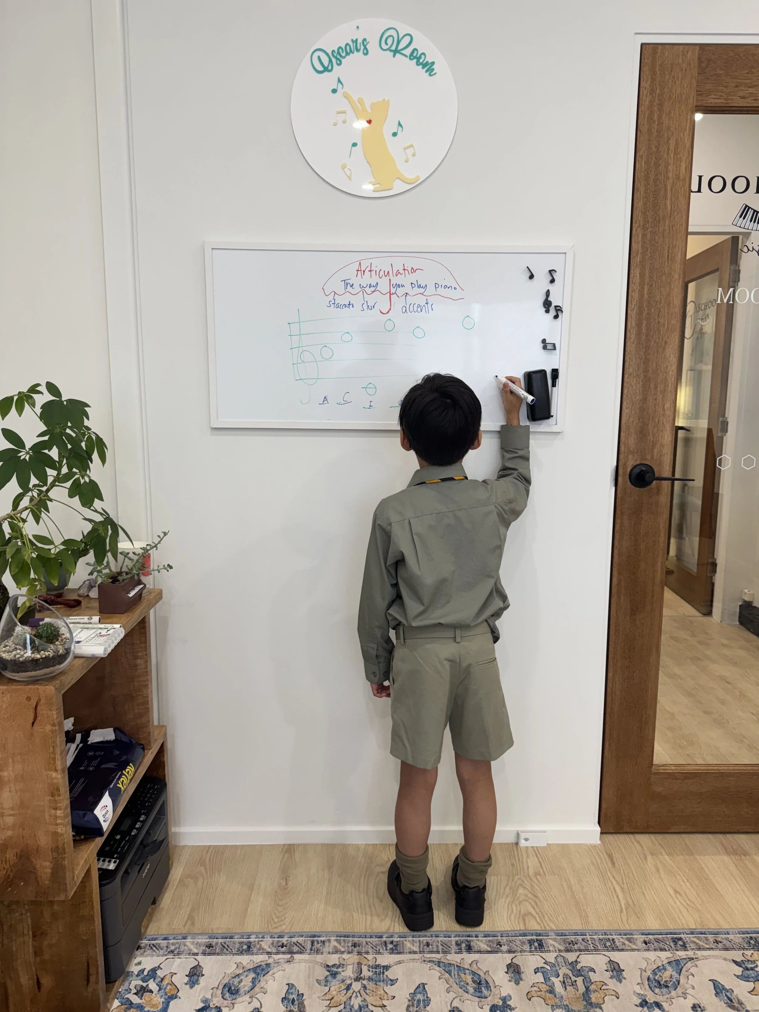 A young boy with black hair, wearing a uniform with khaki shorts, a gray shirt, and hiking boots, stands facing a whiteboard with musical notes and annotations, in a room labeled 'Oscar's Room' with a decorative circular sign above it, and a wooden d