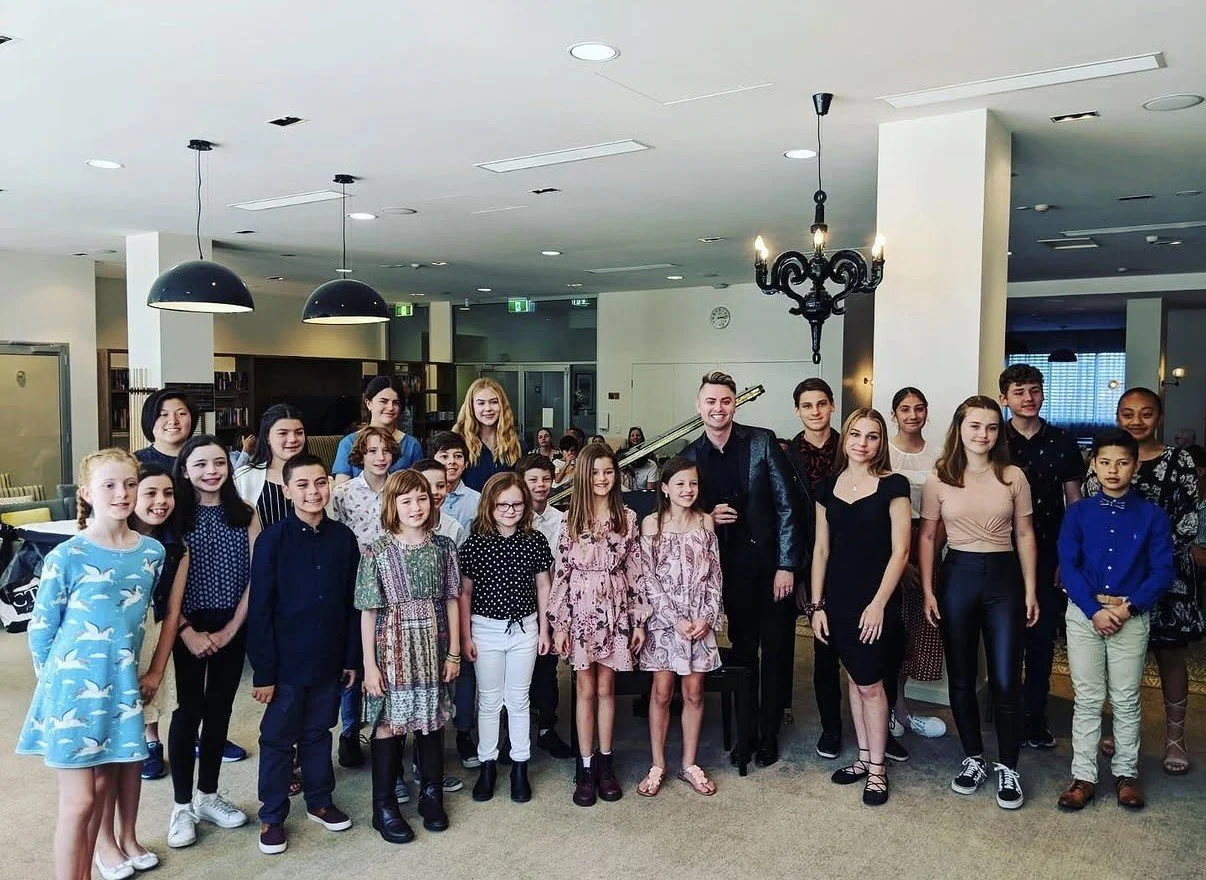 Group of children and teens posing for a photo in an indoor space with a pianist and a chandelier overhead.