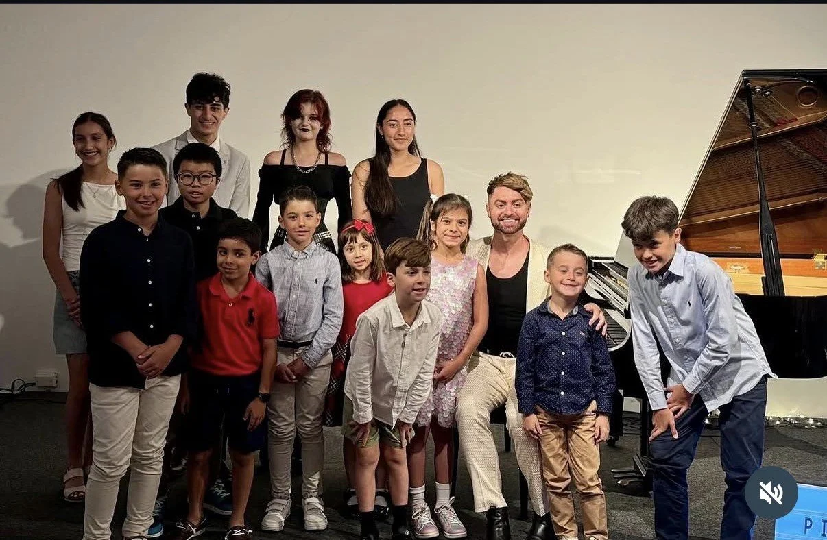 Group of children and adults posing in front of a grand piano on a stage or in a room with plain walls.