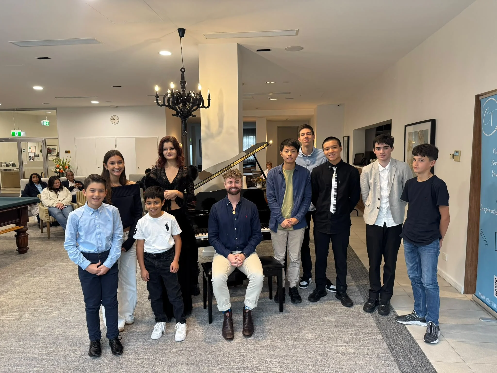 Group of children and adults posing for a photo in an indoor setting with a piano, chandelier, and a blue banner in the background.