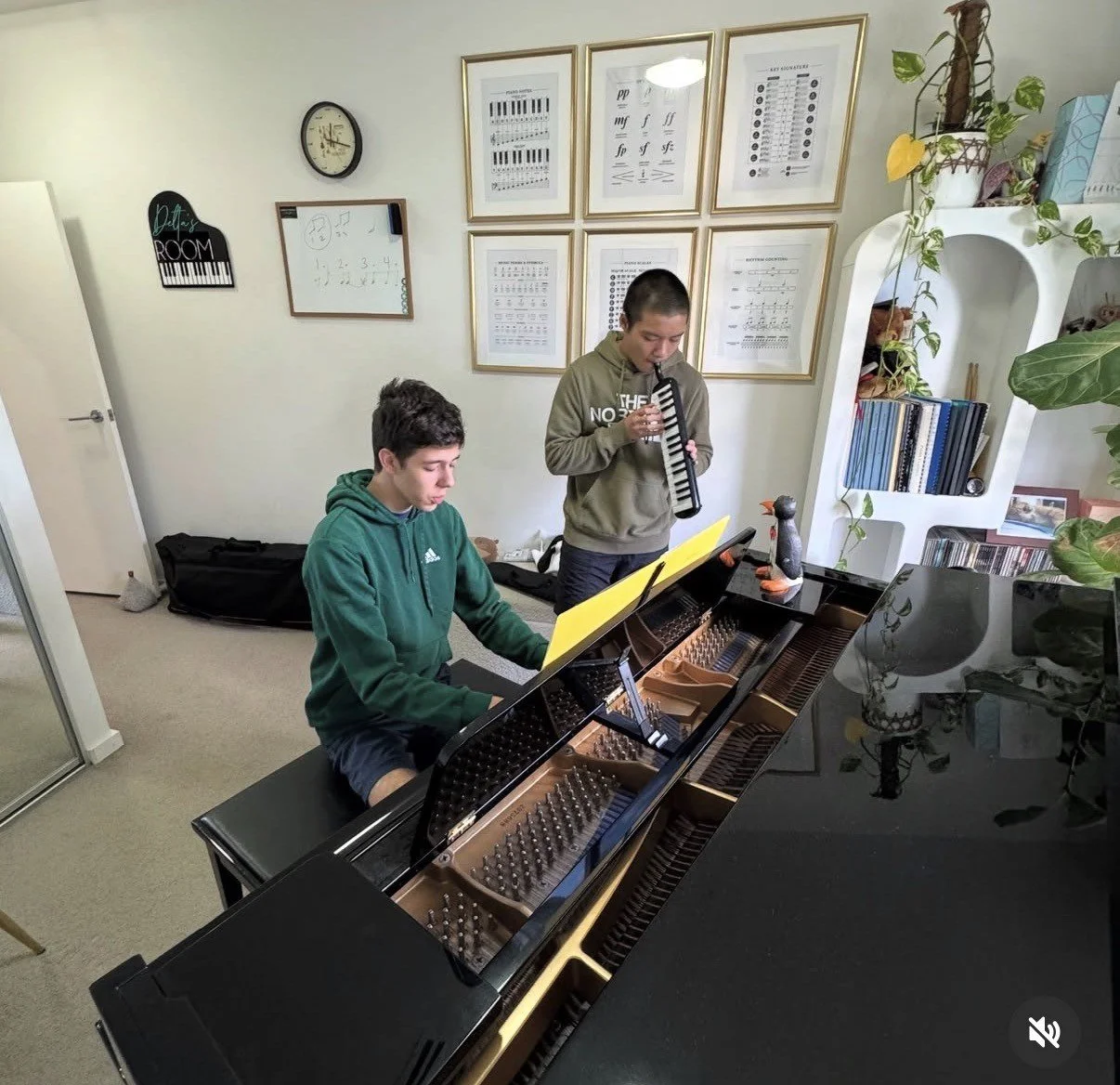 Two young men practicing music in a room with a grand piano, framed music sheets on the wall, a clock, and bookshelves, one playing the piano and the other playing a melodica.