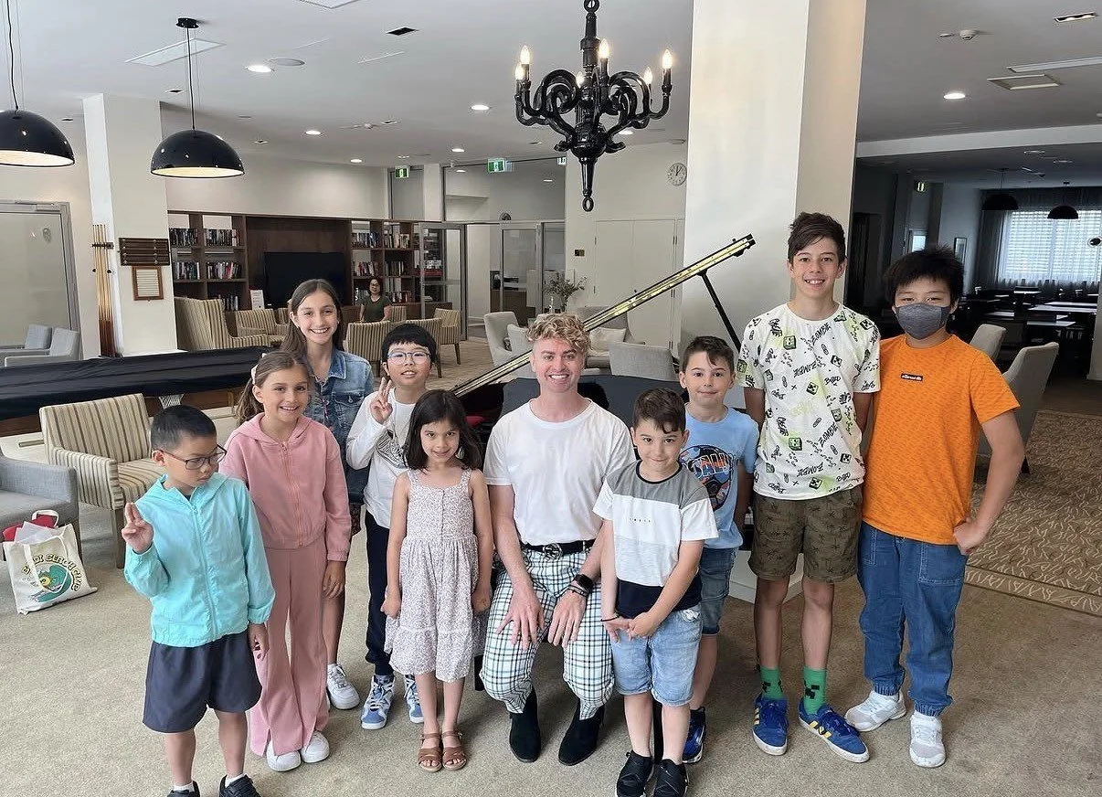 A group of ten children and one adult posing in a room with a grand piano, bookshelves, and chandeliers.
