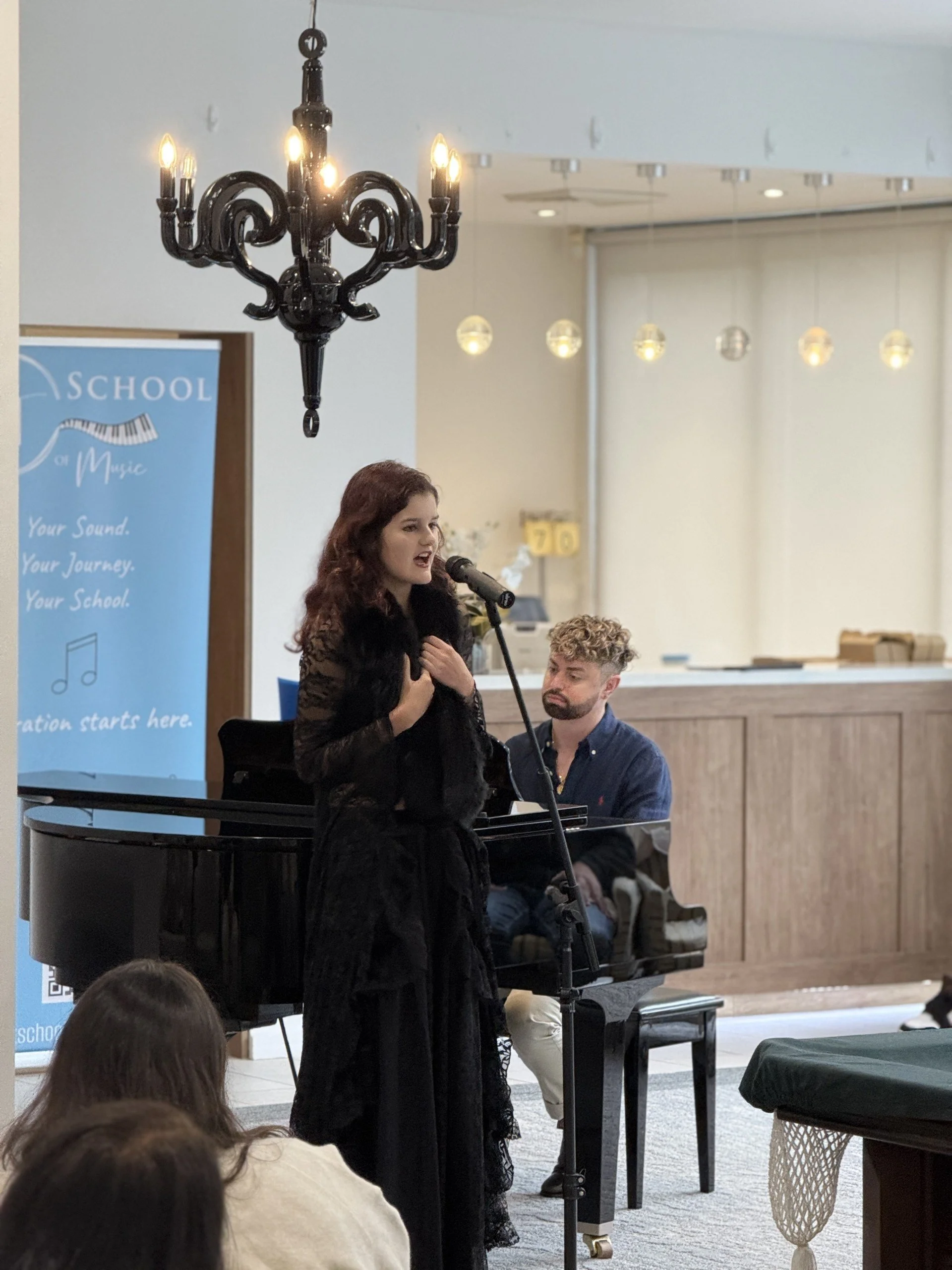 Woman singing with hand on chest while standing in front of a pianist playing a grand piano, in a room with a chandelier and a blue banner.