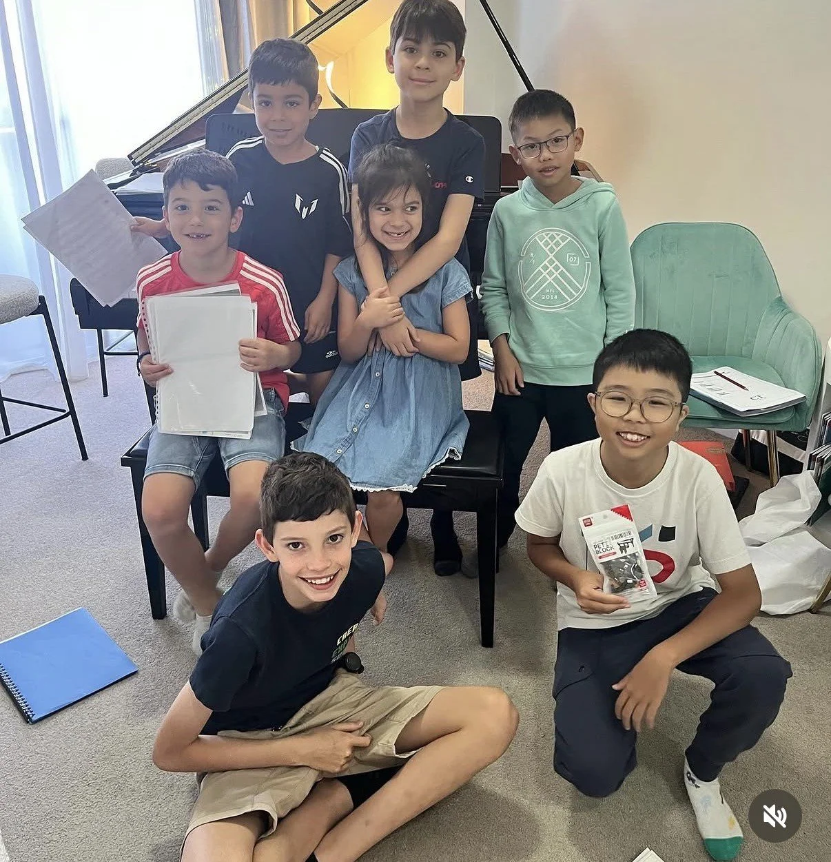 A group of children posing indoors with a grand piano in the background. Some are holding papers or books, and they are smiling or looking at the camera.