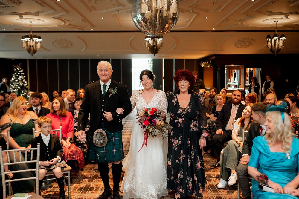 the bride walks down the aisle with her parents as seated guests look on during a wedding ceremony organised by an american destination wedding planner in glasgow scotland