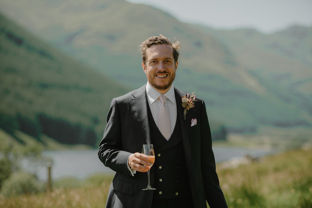 the groom stands in the Scottish countryside holding a glass of champagne with hills and a loch visible in the background before his Monachyle Mhor wedding in Perthshire Scotland