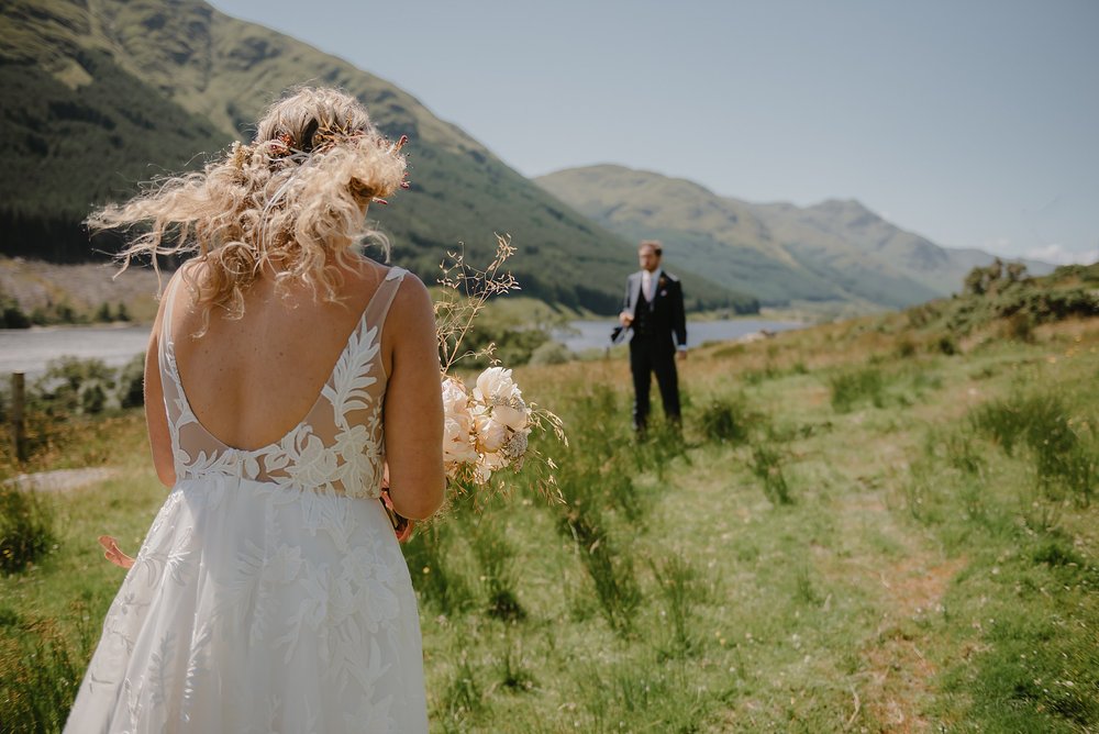 the bride walks towards the groom in the Perthshire countryside before their Monachyle Mhor wedding hills and a loch are visible in the background