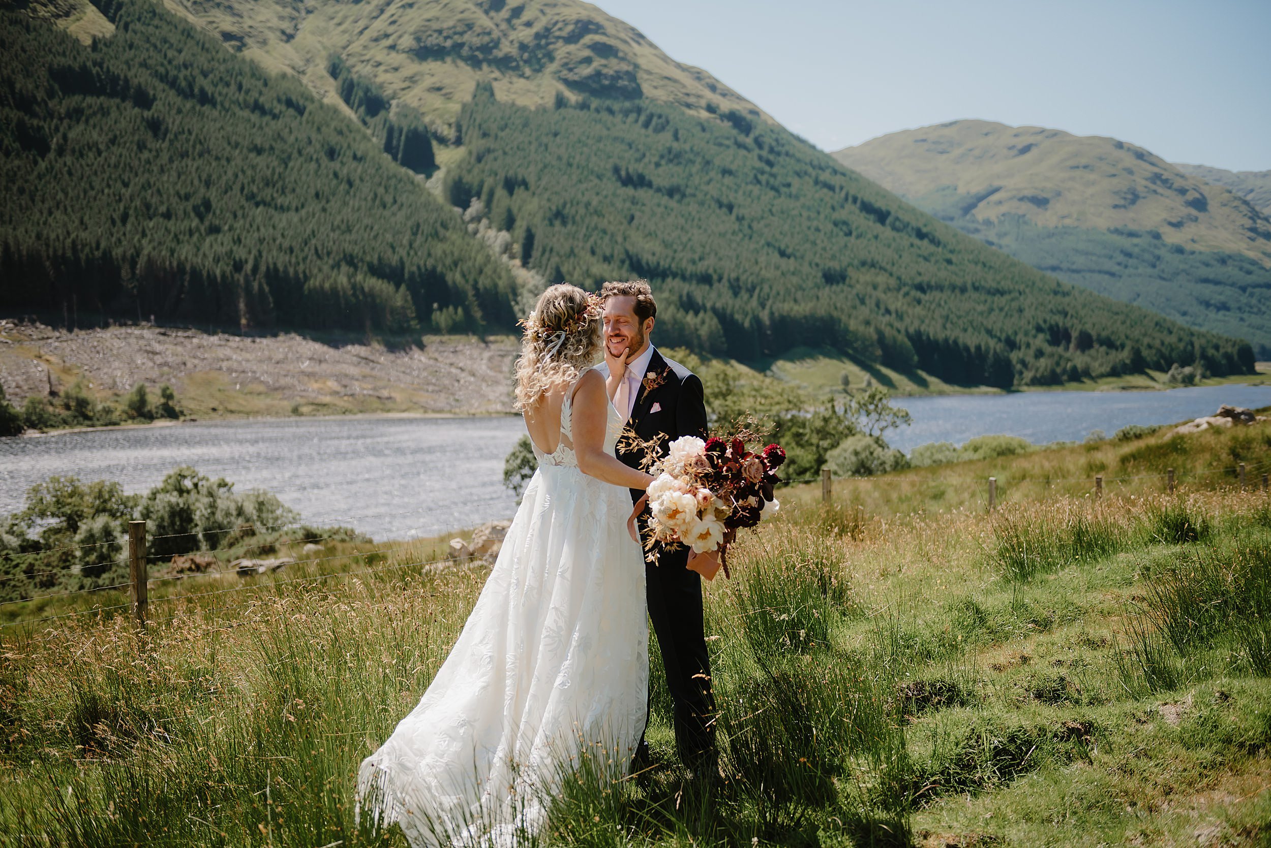 the bride and groom stand face to face in the Perthshire countryside with a loch and hills visible in the background before their Monachyle Mhor wedding in Scotland
