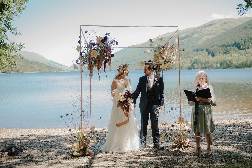 the bride and groom stand beneath the chuppah on the edge of a loch as their celebrant conducts their Monachyle Mhor wedding ceremony