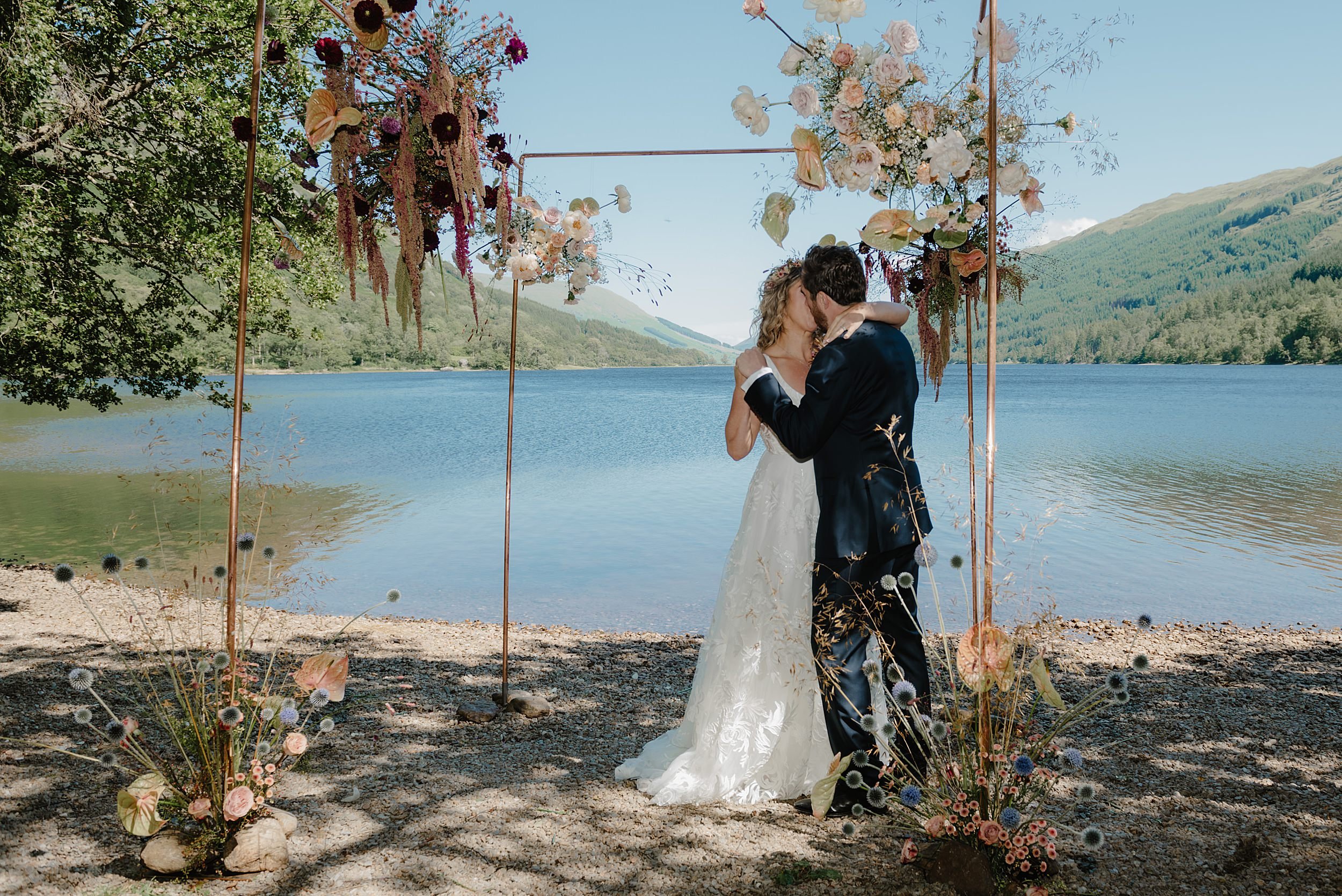 the bride and groom kiss beneath the chuppah on the edge of a loch during their Monachyle Mhor wedding ceremony in Perthshire Scotland