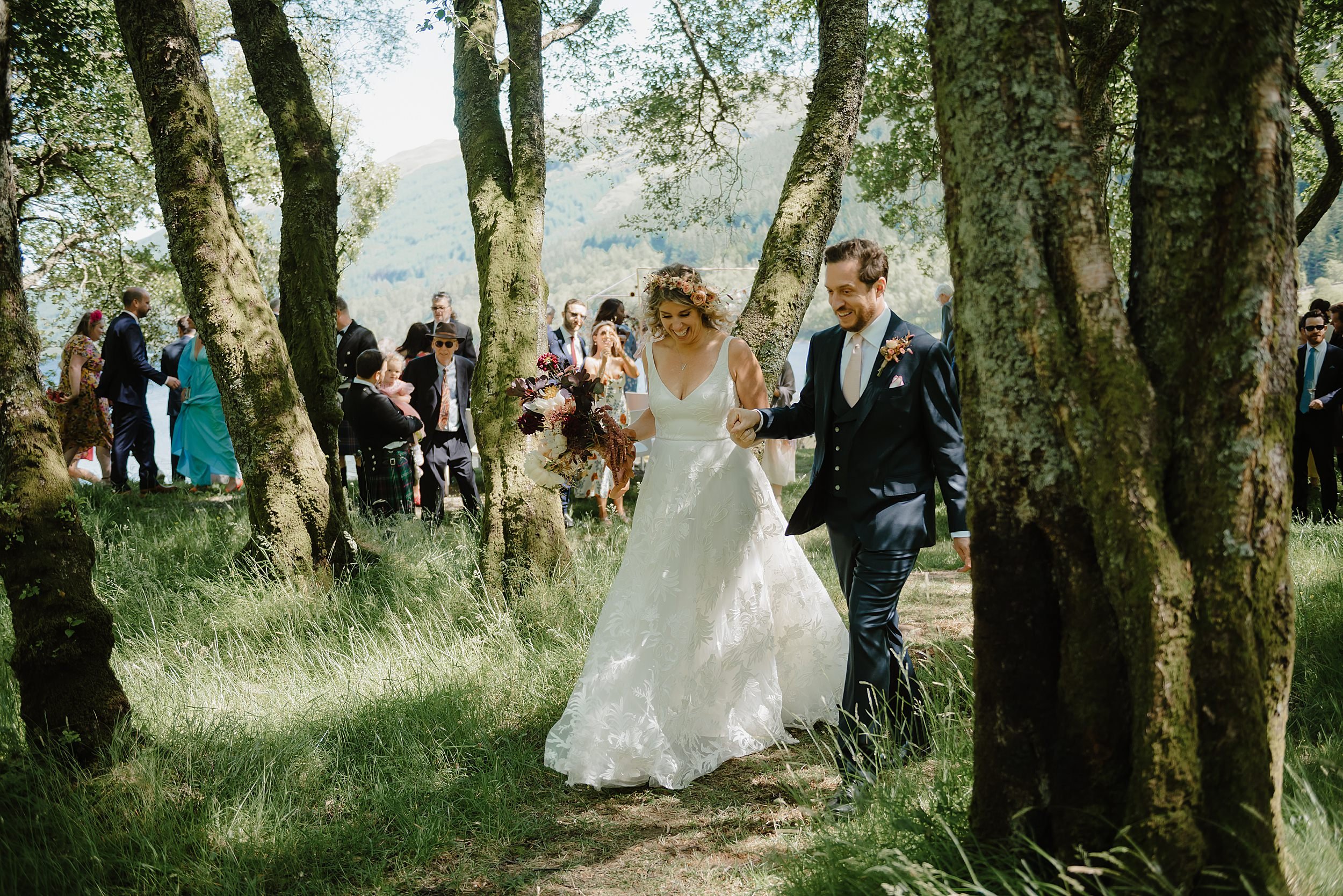 the bride and groom walk through a wooded area followed by their guests after their Monachyle Mhor wedding ceremony in Perthshire in Scotland