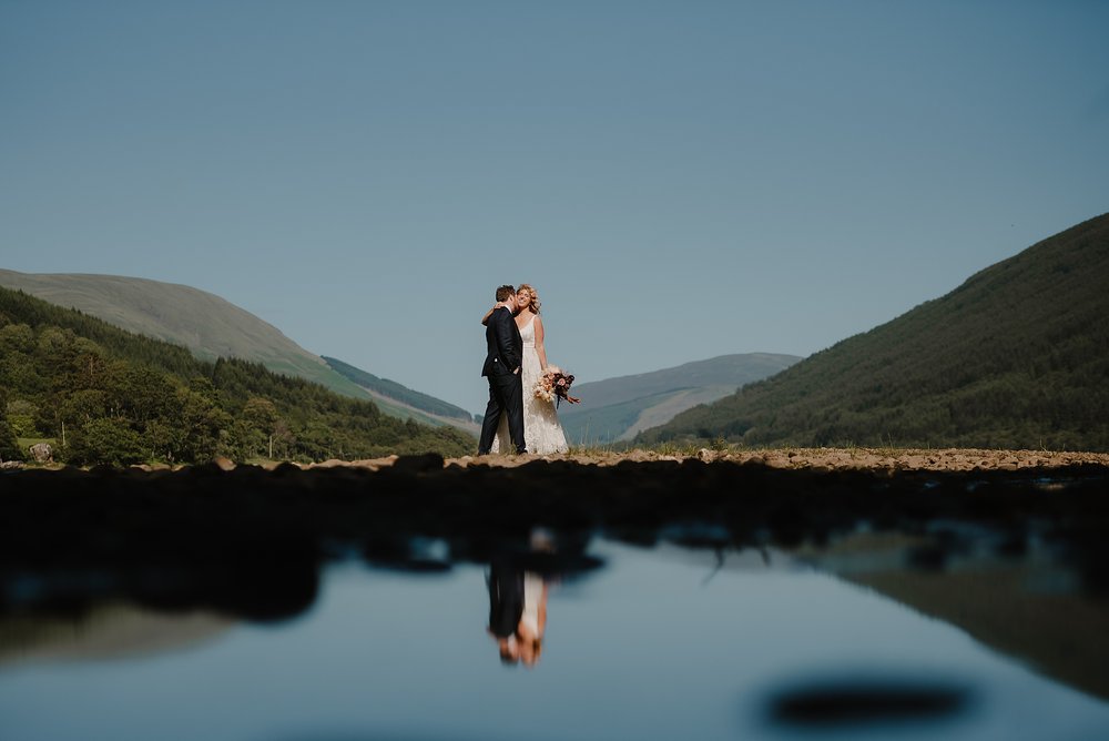 the bride and groom embrace standing at the edge of a loch near Monachyle Mhor wedding venue in Perthshire in Scotland