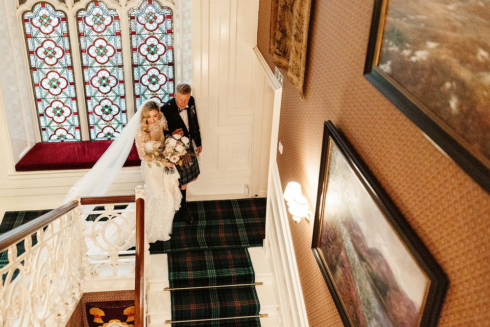 bride and her father walking down the stairs inside drumtochty castle on her way to her wedding ceremony planned by Scottish destination wedding planner