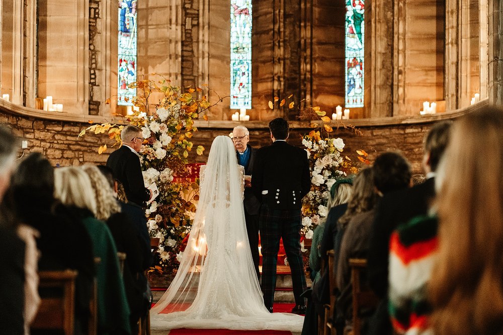 bride and groom standing at Scottish church alter with candles and autumnal fresh flowers during their wedding ceremony by Scottish destination wedding planner