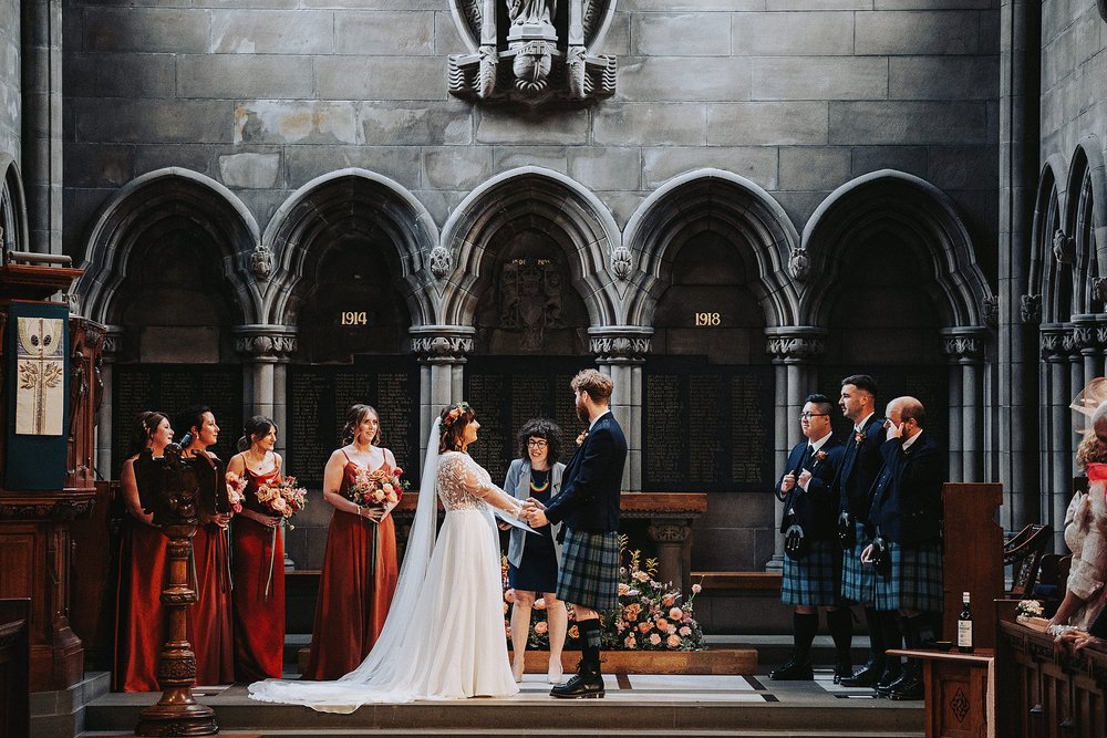 bride and groom getting married and smiling at each other standing at the altar of glasgow university memorial chapel before the couple's diy wedding venue scotland reception at barrowlands