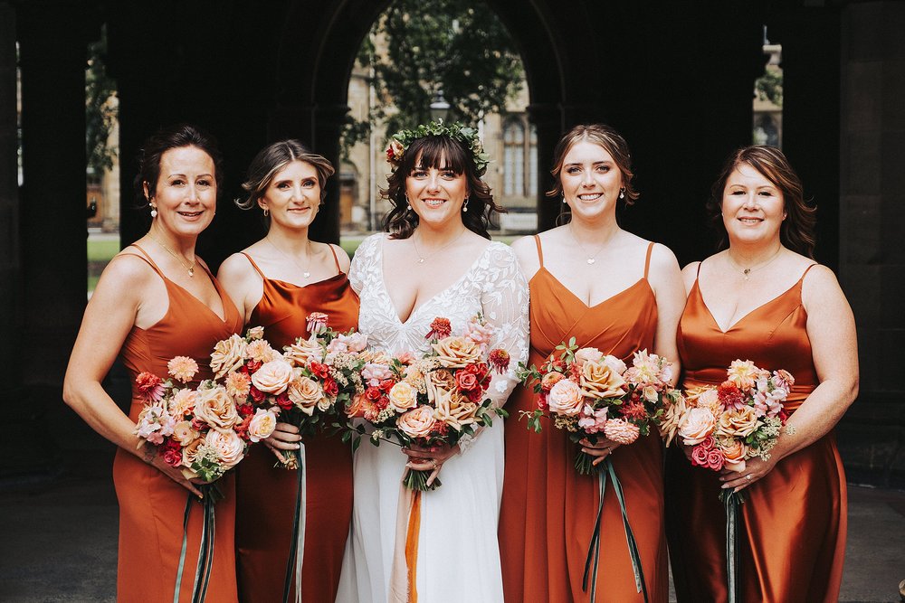 bride standing with her bridesmaids wearing burnt orange silk bridesmaid dresses standing out glasgow university memorial chapel before the couple's diy wedding venue scotland reception at barrowlands