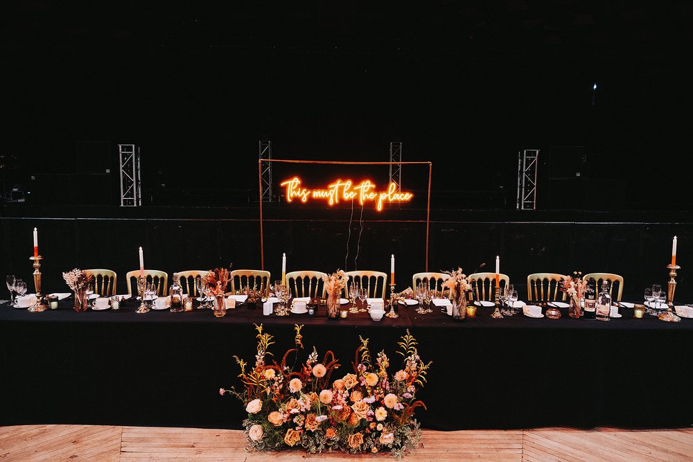 inside barrowlands which is a diy wedding venue scotland showing the top table with a black table cloth autumnal flowers and an orange neon sign saying this must be the place