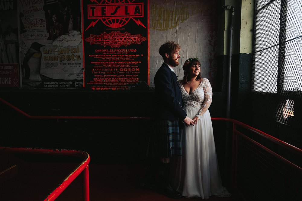 bride and groom standing on the stairs in front of a gig poster and looking out the window inside barrowlands which is a diy wedding venue scotland