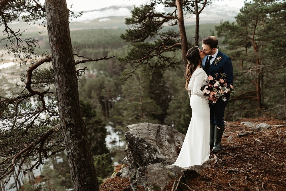 bride and groom standing amongst a Scottish pine forest overlooking a loch on a snowy winter's day during their their elopement by Scottish highlands destination wedding planner