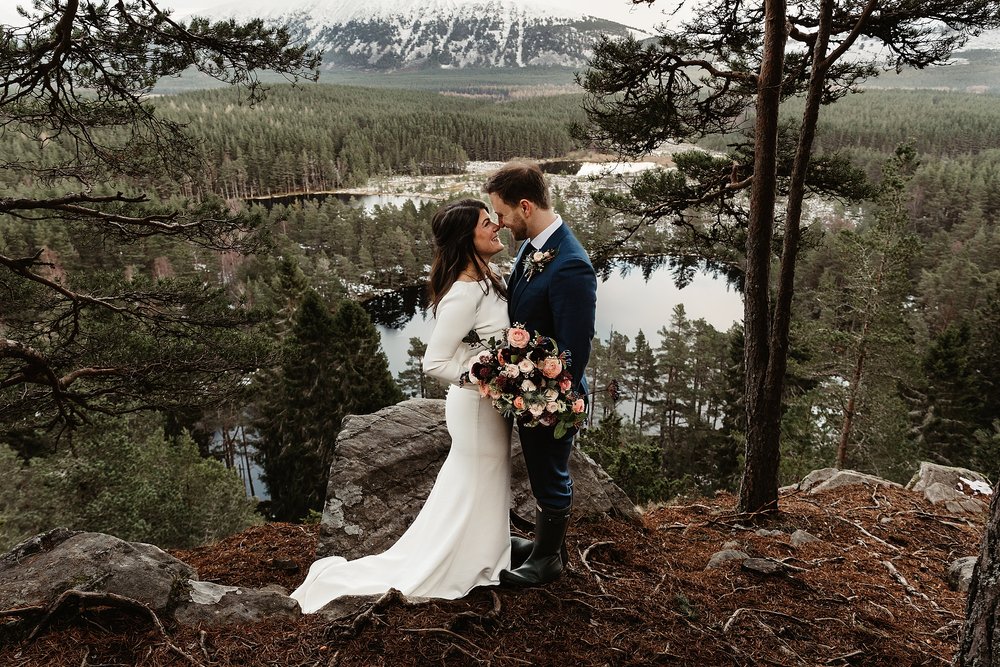 bride and groom kissing in a cairngorm pine forest overlooking a loch on a snowy day during their their elopement by Scottish highlands destination wedding planner