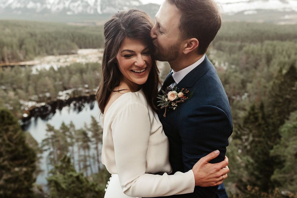 groom hugging and kissing his bride as they stand above a loch in the cairngorms on a winters day for their outdoor Scottish elopement ceremony