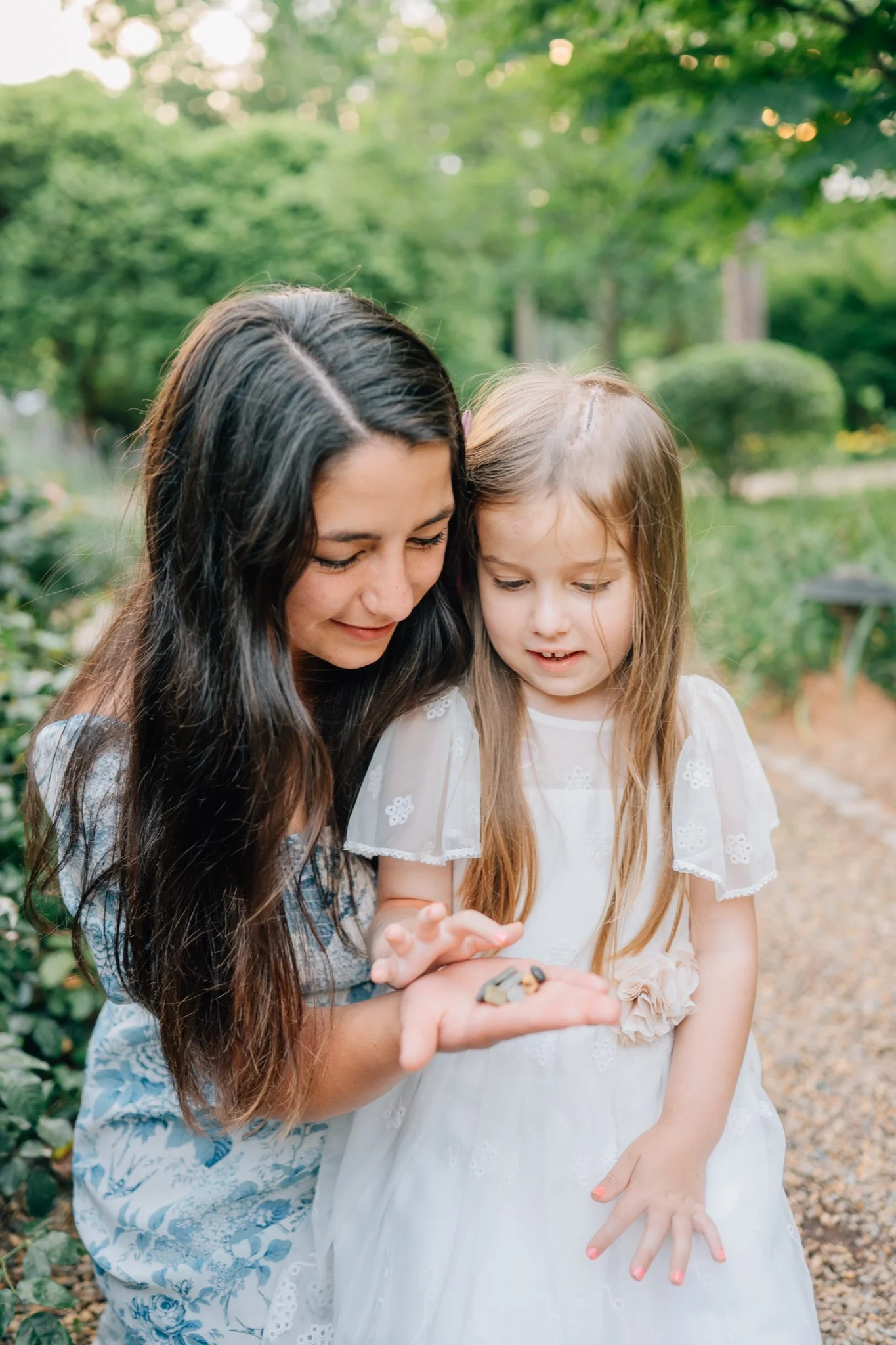    A mother kneels and kisses the forehead of her older daughter while carrying her younger daughter on her shoulders in a rose garden at sunset.     Mother and young daughter smiling at each other in front of a peaceful lake, with sunlight filtering