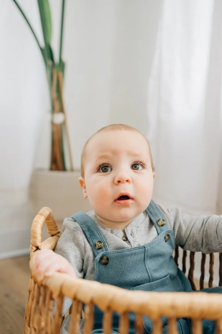  Parents sitting close with their baby in a bright Greenville natural light studio, soft neutral tones and joyful expressions.  Close-up profile of a one-year-old baby looking toward window light in a calm Greenville studio.  Father lifting his baby 