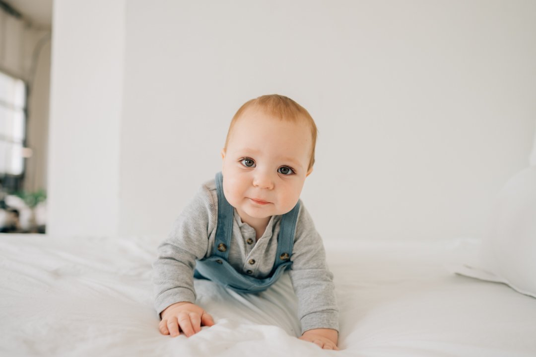  Parents sitting close with their baby in a bright Greenville natural light studio, soft neutral tones and joyful expressions.  Close-up profile of a one-year-old baby looking toward window light in a calm Greenville studio.  Father lifting his baby 