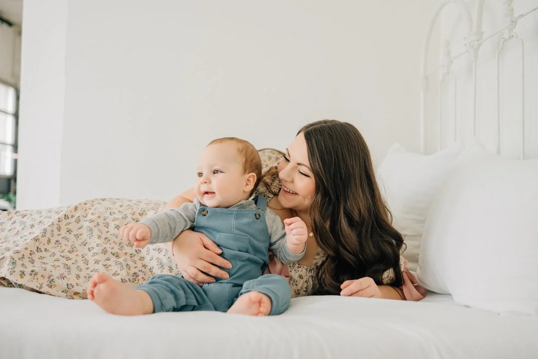  Parents sitting close with their baby in a bright Greenville natural light studio, soft neutral tones and joyful expressions.  Close-up profile of a one-year-old baby looking toward window light in a calm Greenville studio.  Father lifting his baby 