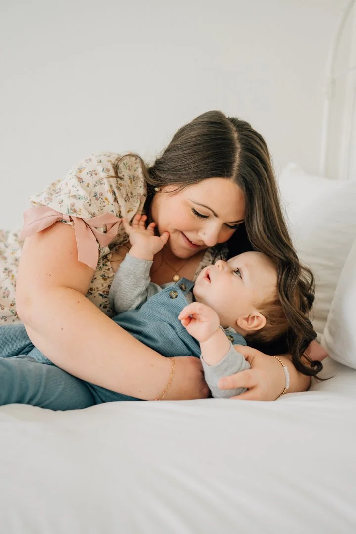  Parents sitting close with their baby in a bright Greenville natural light studio, soft neutral tones and joyful expressions.  Close-up profile of a one-year-old baby looking toward window light in a calm Greenville studio.  Father lifting his baby 