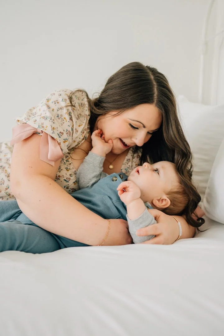  Parents sitting close with their baby in a bright Greenville natural light studio, soft neutral tones and joyful expressions.  Close-up profile of a one-year-old baby looking toward window light in a calm Greenville studio.  Father lifting his baby 
