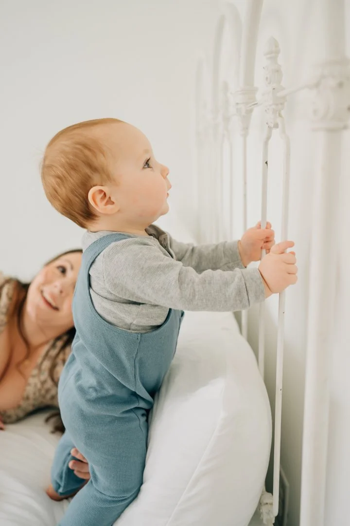  Parents sitting close with their baby in a bright Greenville natural light studio, soft neutral tones and joyful expressions.  Close-up profile of a one-year-old baby looking toward window light in a calm Greenville studio.  Father lifting his baby 